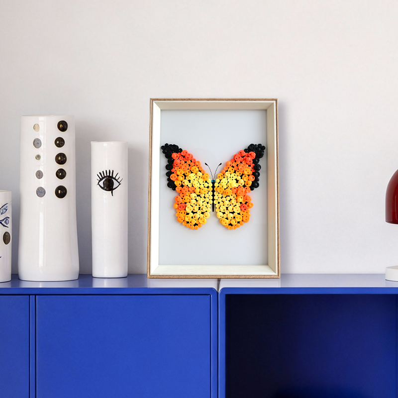 Orange butterfly button art in a frame on a shelf with other decorative items.
